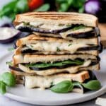 Two eggplant paninis stacked on top of each other on a white plate. There are basil leaves scattered around.