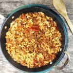 Aerial view of prepared American Chop Suey in a large skillet sitting on a wooden background. There is a light colored wooden spoon to the upper right of the pan.