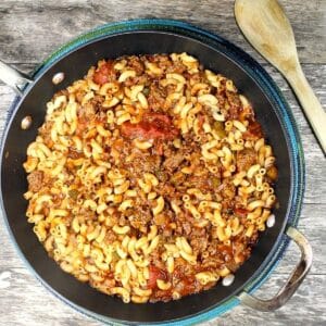 Aerial view of prepared American Chop Suey in a large skillet sitting on a wooden background. There is a light colored wooden spoon to the upper right of the pan.