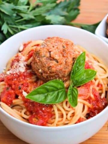 close up of pasta in a white bowl with red sauce and a meatball on top. There is a basil leaf propped next to meatball.