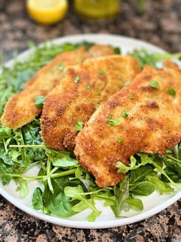 Three fried chicken cutlets on a bed of greens sitting on a white dish. The dish is on a black and brown counter with a lemon behind it.