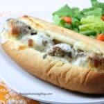 Prepared meatball sub on a white plate. It is close up shot with a green salad to the right of the sub.