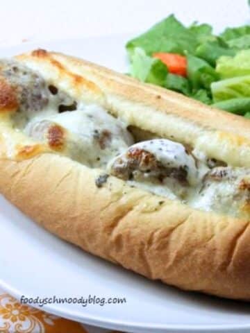 Prepared meatball sub on a white plate. It is close up shot with a green salad to the right of the sub.