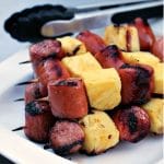 skewers with smoked kielbasa and pineapple on plate with tongs behind the plate