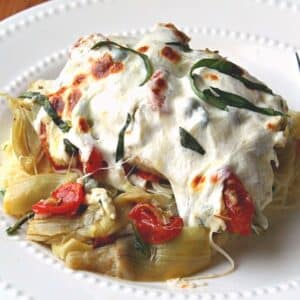 close up of cheese covered chicken with artichokes and tomatoes on a white dish.