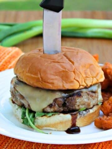 Burger in a bun on plate, dripping with glaze. There is a knife vertically through the burger.