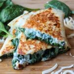 Cooked spinach quesadilla triangles piled atop each other. They are piled on a wooden cutting board.