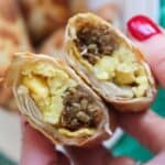 A close up of a hand holding two halves of an egg roll showing the eggs and sausage inside.