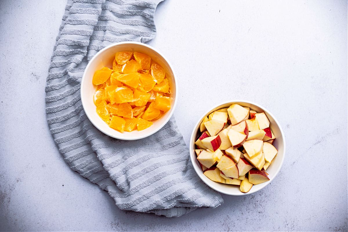 chopped up apples in a bowl next to a bowl of chopped oranges with a grey towel next to them
