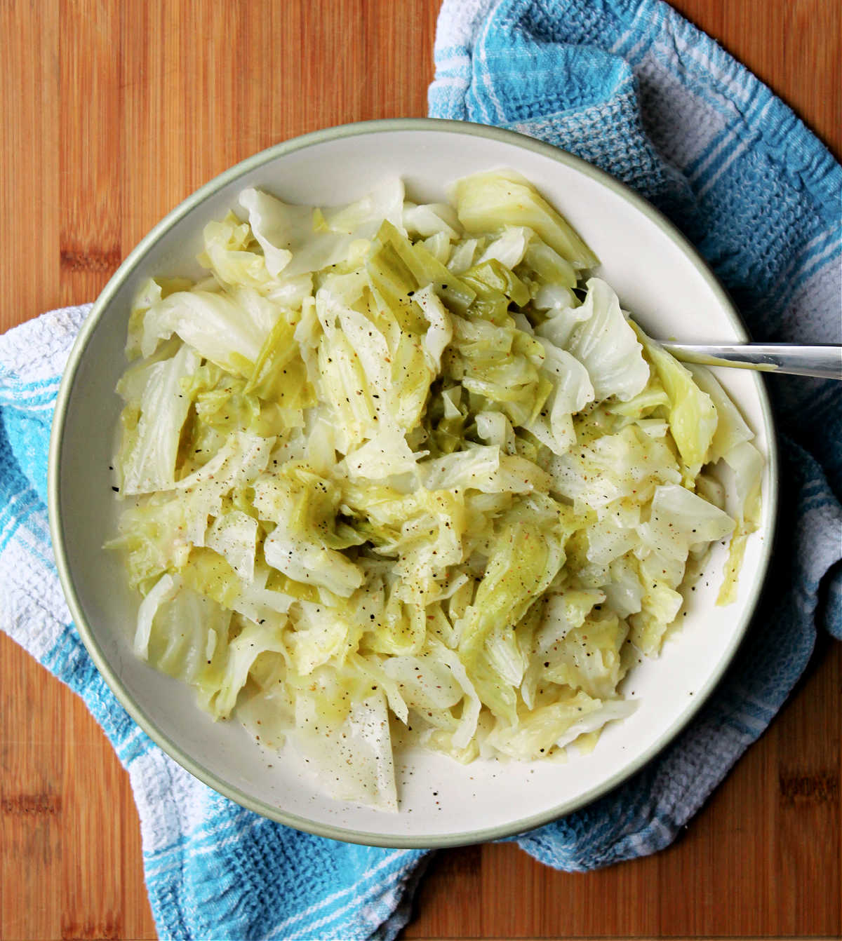 cooked cabbage in serving bowl with spoon in bowl. Bowl is sitting atop a blue and white dish towel.