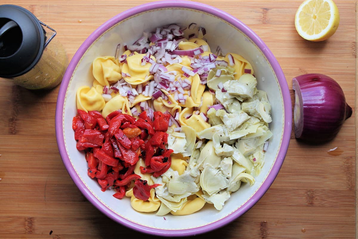 white bowl with purple border. inside bowl is the cooked tortellini, chopped roasted red pepper, chopped artichoke hearts and diced red onion. To the right of the bowl is half of a red onion and half of a lemon.