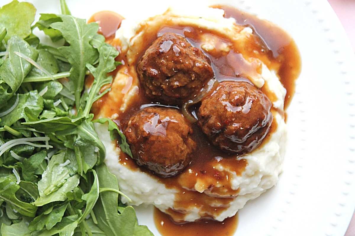 An aerial view of 3 meatballs atop a pile of mashed potatoes, smothered in gravy on a white plate. There is an arugula salad to the left of the mashed potatoes.
