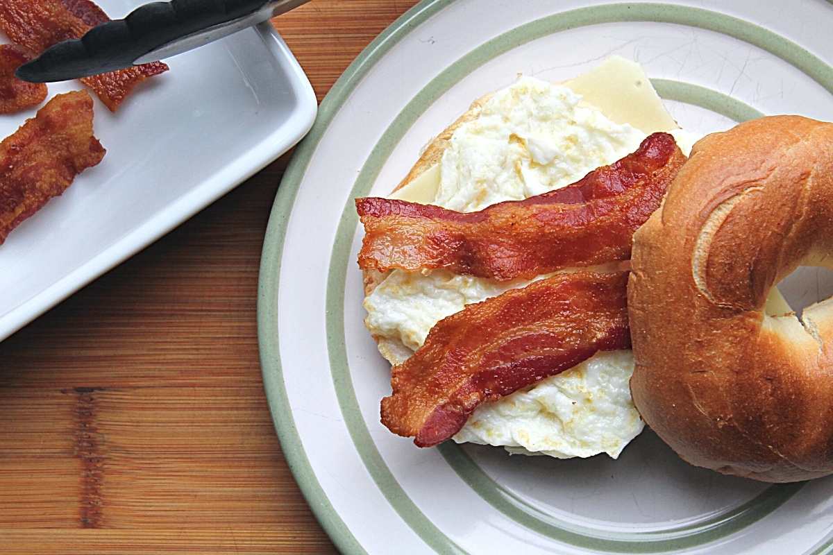 Bacon is layered on a bagel and egg sandwich on a white and green plate. The edge of a white bowl with cooked bacon is just to the left of the plate.