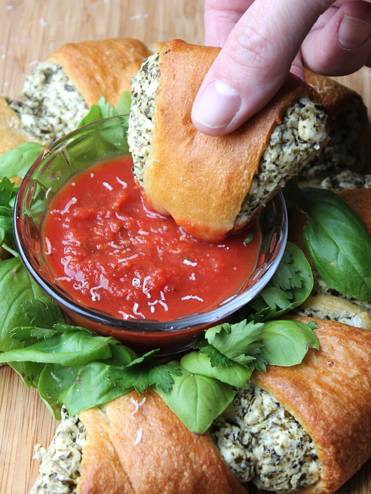 A hand is holding a stuffed crescent roll, dipping it in marinara sauce which is in the center of a crescent roll wreath.