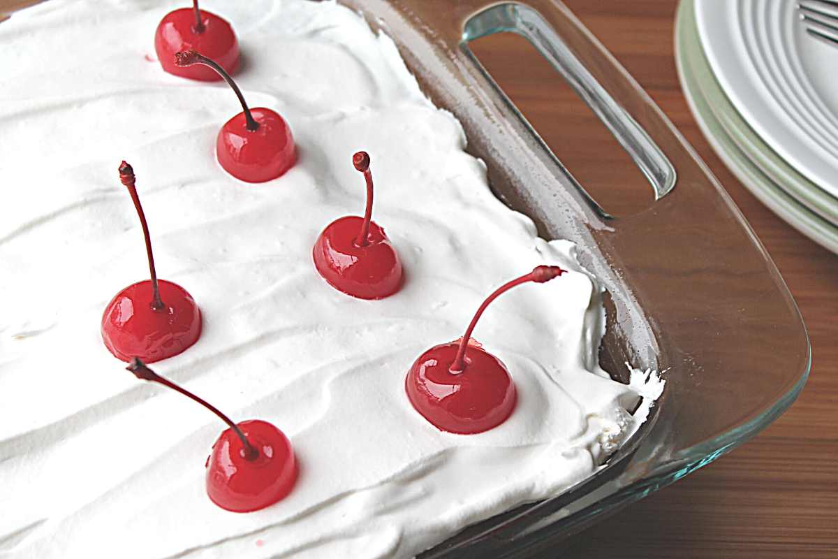 Corner of cake pan is showing prepared cake with whipped topping and cherries on top. There are dishes to the side of the cake pan.
