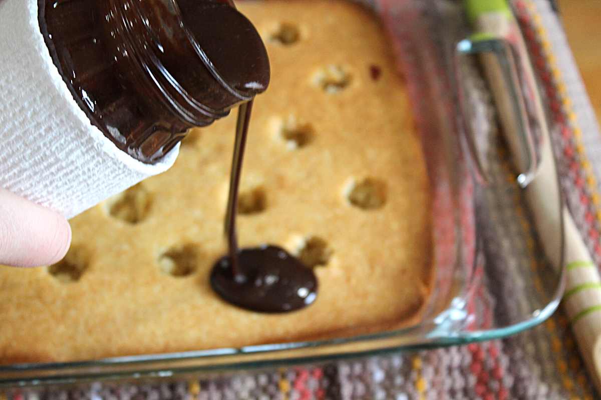 Hot fudge sauce being poured onto vanilla cake in pan. The cake has holes poked all over it.