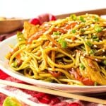 cooked lo mein served on a white dish. There are chopticks in front of dish and it is all on a red and white tablecloth.