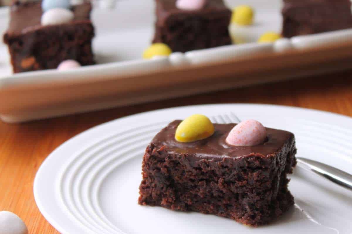 A brownie, topped with two candy eggs on a white plate. There is a white platter in the background with more brownies on it.