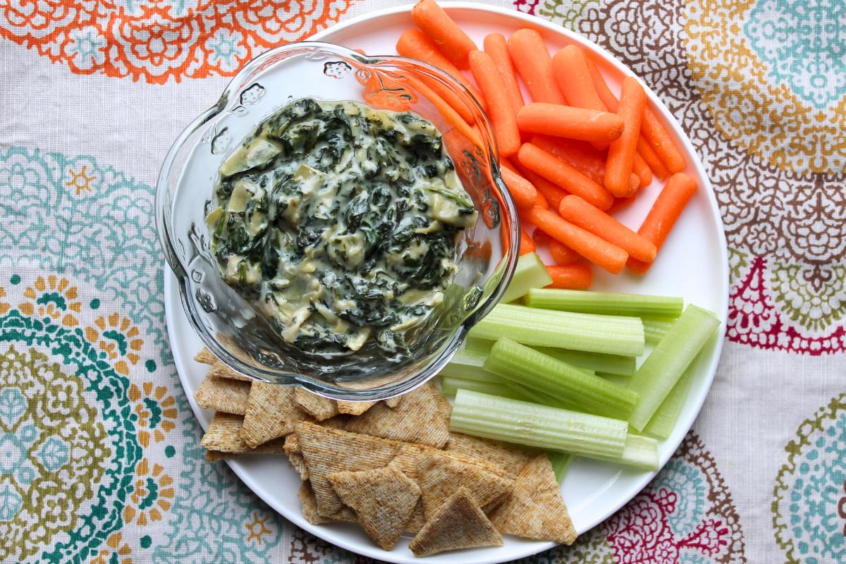 A white plate sits on top of a mutilcolored tablecloth. On the plate is a bowl of prepared dip, baby carrots, celery and crackers.