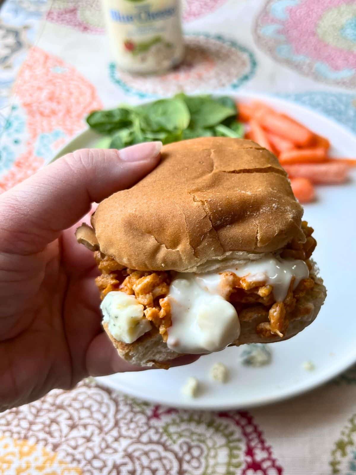 A hand holding a buffalo chicken slider with blue cheese dressing dripping. In the background is a white plate with carrot sticks.