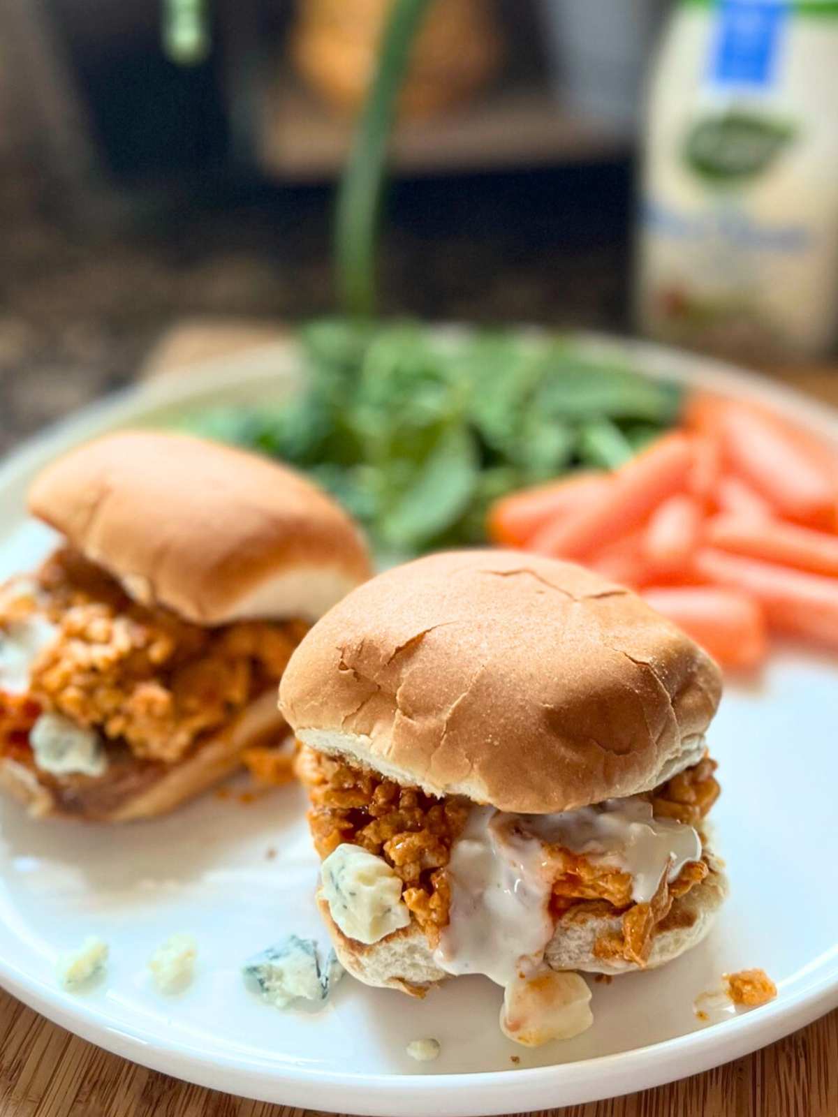 A white plate with two chicken sliders on it. There is also carrot and salad blurred behind the sliders. There is dressing dripping from the sliders.