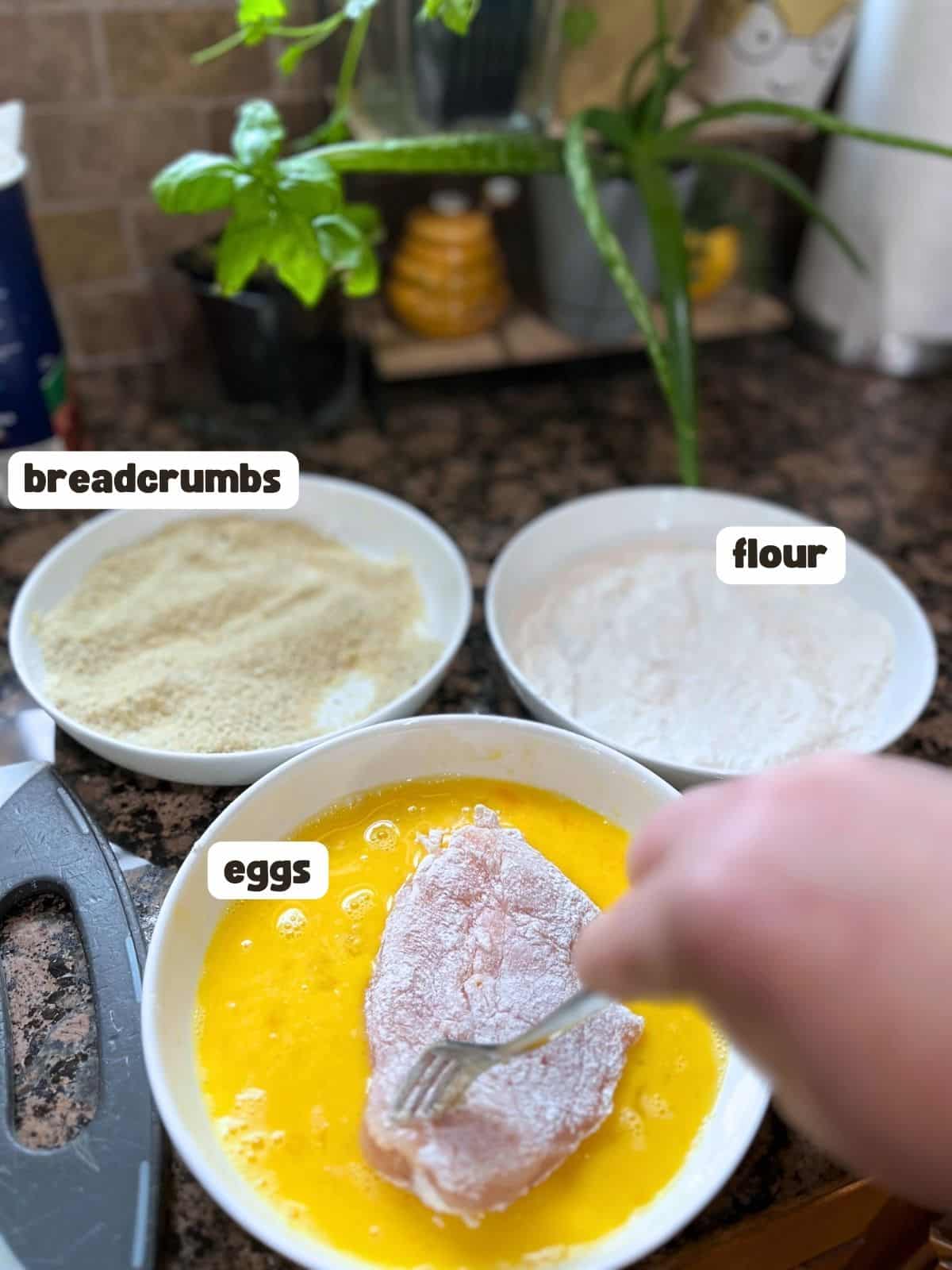 Three shallow white bowls. One filled with breadcrumbs, one with flour and one with eggs. There is a hand dripping coated chicken into the eggs.