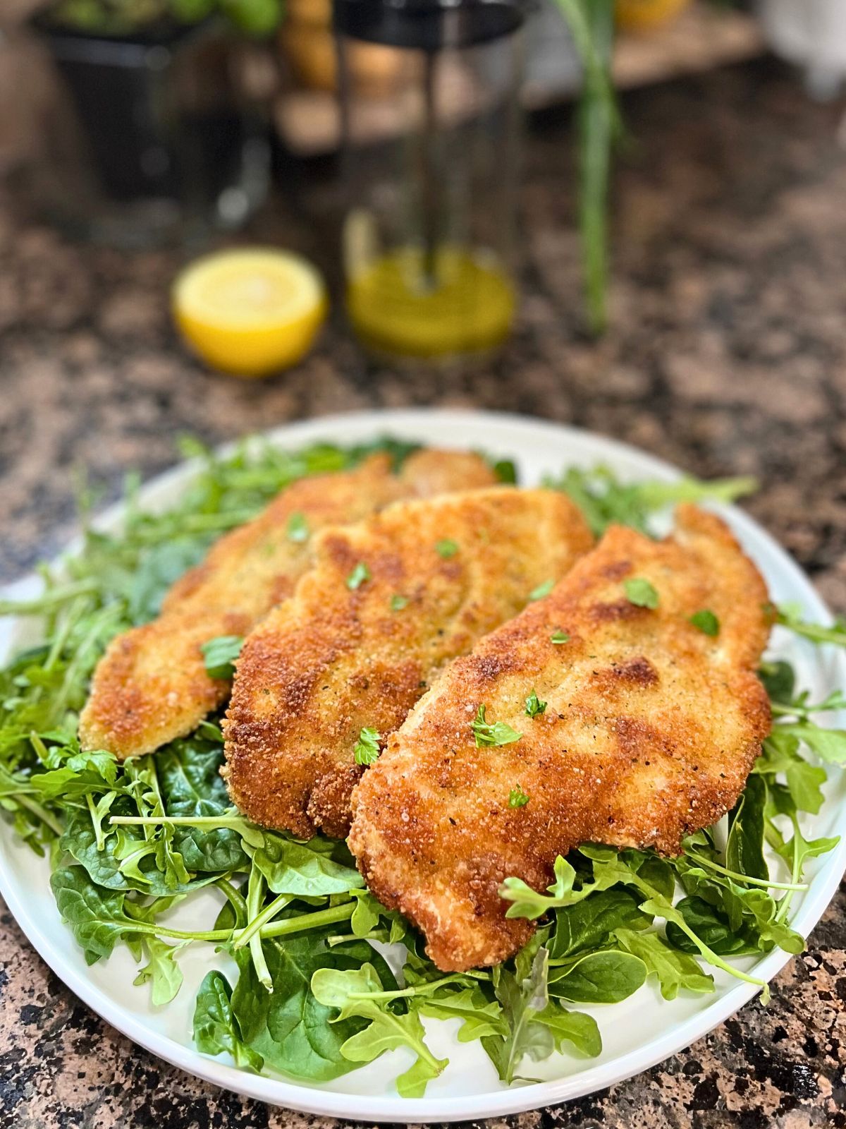 3 Crispy Chicken cutlets are on a bed of arugula greens which is on a white serving plate. Blurred in the back is a half lemon and a jar of dressing.