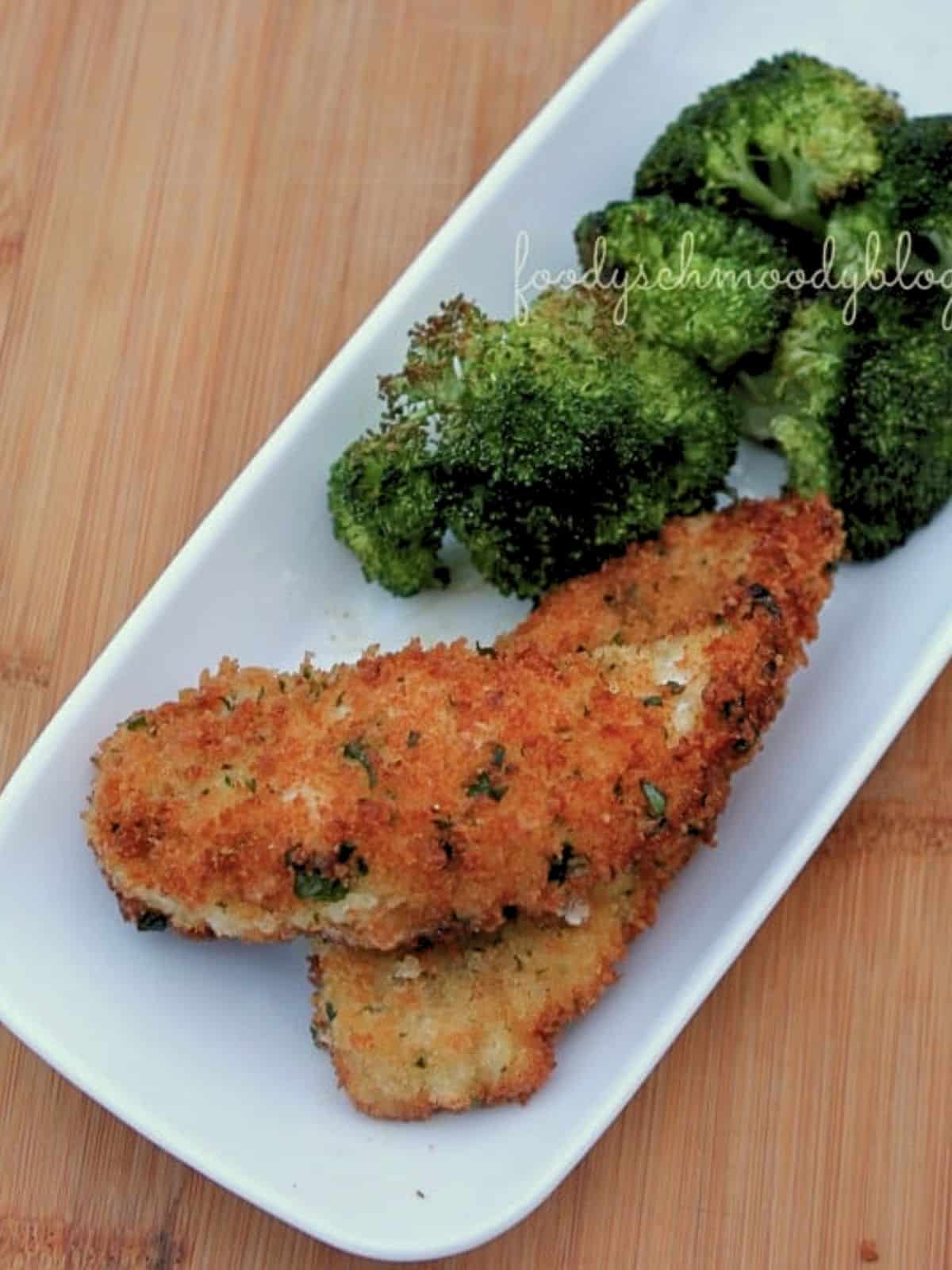 Two chicken tenders on a long white plate with broccoli next to it.