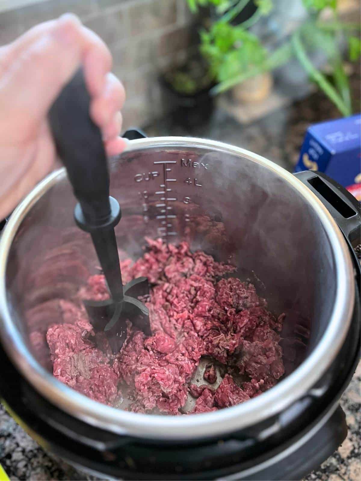Ground beef being cooked in the instant pot. There is a hand using a utensil to break up beef.