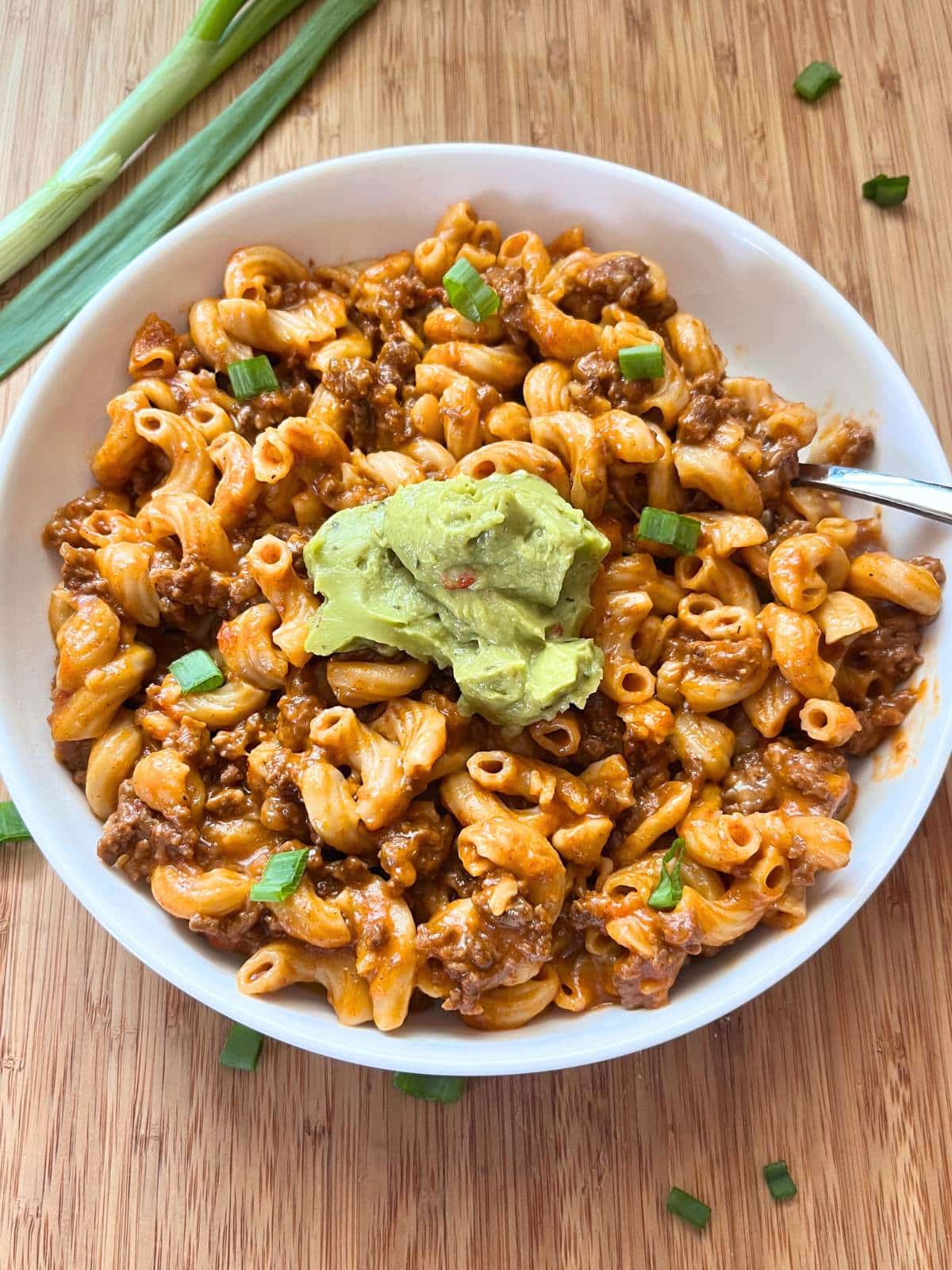 A close up overhead view of cooked taco pasta in a white bowl. The pasta is topped with a dollop of guacamole. The bowl is on a wooden cutting board with a green onion to the top left.