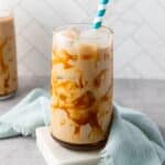 caramel iced coffee in a large glass. The glass is on a white coaster and there is a light blue kitchen towel draped behind and to the side of it.