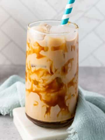 caramel iced coffee in a large glass. The glass is on a white coaster and there is a light blue kitchen towel draped behind and to the side of it.