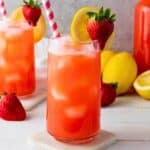 A tall glass with prepared strawberry lemonade in it. There is a second filled glass to the left and lemons just behind them both.