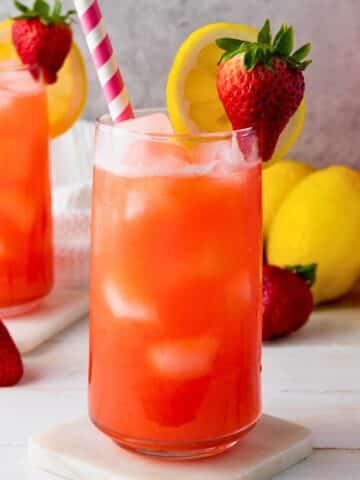A tall glass with prepared strawberry lemonade in it. There is a second filled glass to the left and lemons just behind them both.