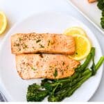 Two cooked fillets of salmon on a white plate with two lemon slices and cooked broccolini.