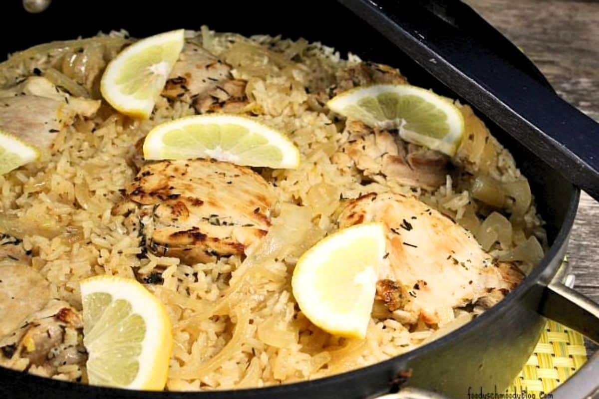 Prepared chicken and rice topped with lemon slices in a skillet. the skillet is on a yellow placemat on a wooden surface.