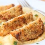Sliced cooked breaded pork chop sitting on top of cooked polenta in a white plate. The view is up close with nothing else visible from background.