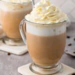 A hot vanilla latte topped with whipped cream. The latte is served in a glass mug as to see the layers. There is a second latte to the left background.