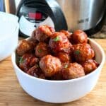 Prepared cranberry meatballs in a white serving bowl. The bowl of meatballs sits on a wooden cutting board with a slow cooker in the background.