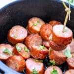 A close up of cooked sliced kielbasa in an air fryer basket. There is a toothpick skewer that is lifting two slices out of the basket.