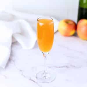 A champagne flute holding a prepared mimosa. There is a white napkin to the back left and apples blurred out on the back right.