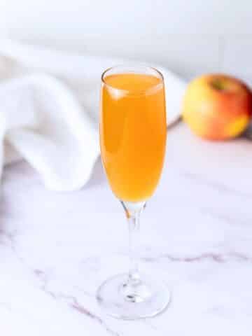 A champagne flute holding a prepared mimosa. There is a white napkin to the back left and apples blurred out on the back right.