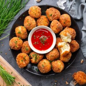 A black plate filled with prepared mashed potato balls. In the center of the plate is a small white bowl of marinara for dipping. The plate is resting on a slate background and there are herbs scattered around it.