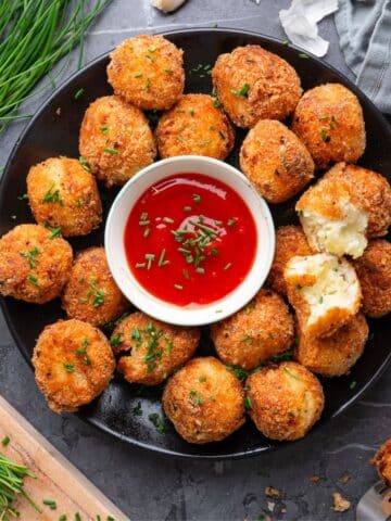 A black plate filled with prepared mashed potato balls. In the center of the plate is a small white bowl of marinara for dipping. The plate is resting on a slate background and there are herbs scattered around it.