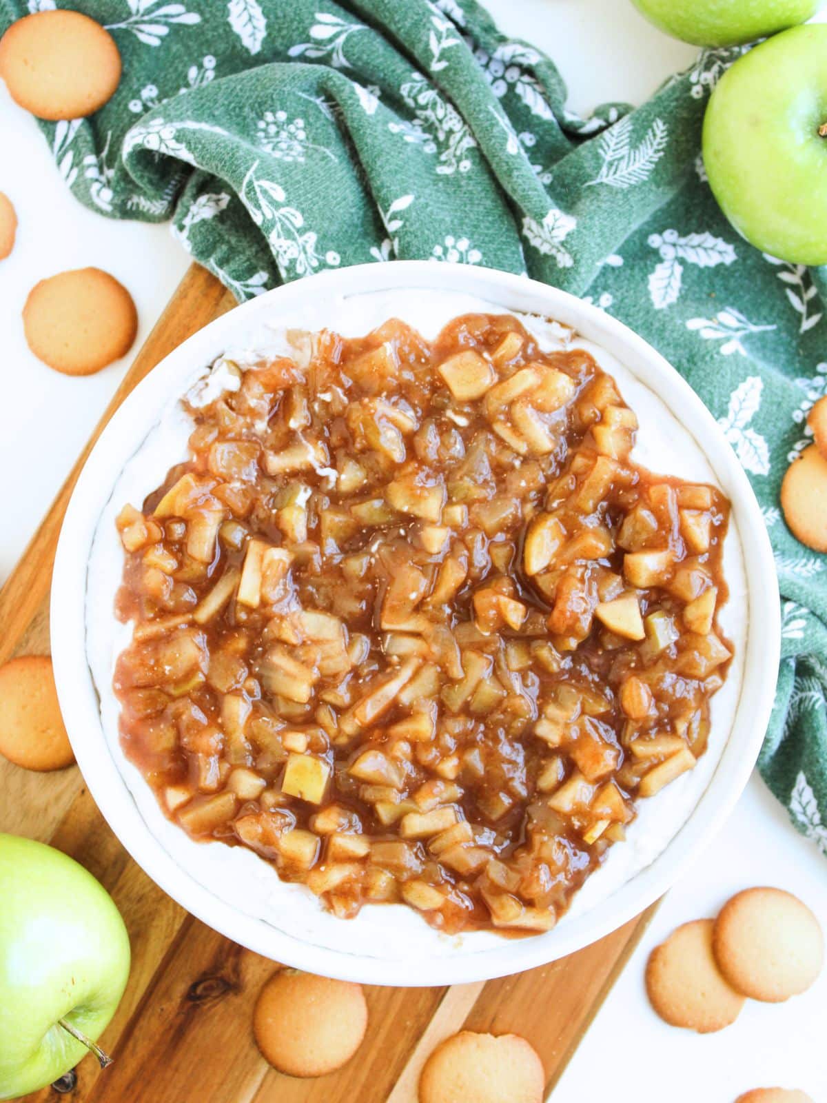 Apple pie dip being served on a white plate which is sitting on a wooden board. There are apples and wafter cookies scattered around the dip.