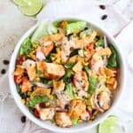 An aerial view of the completed salad with dressing drizzled on top. There are slices of limes scattered around the salad which is in a white bowl.