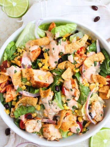 An aerial view of the completed salad with dressing drizzled on top. There are slices of limes scattered around the salad which is in a white bowl.