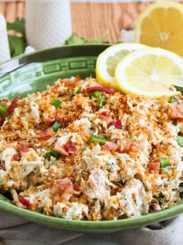 A close up of a green serving bowl full of the prepared chicken salad topped with panko crumbs. Salt and pepper shaker is positioned behind bowl.