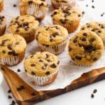 A wooden cutting board covered in parchment paper with several prepared muffins scattered on it. there are chocolate chips scattered all around the board.