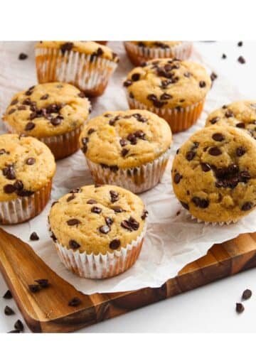 Many prepared muffins on a parchment paper lined wooden cutting board. There are chocolate chips scattered all around.